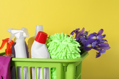 Photo of Spring cleaning. Basket with detergents, flowers and tools on yellow background, closeup