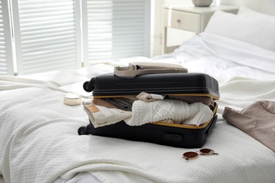 Open suitcase full of clothes, jacket and fashionable accessories on bed in room