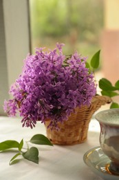 Beautiful lilac flowers in wicker basket and cup of hot coffee on window sill indoors, closeup