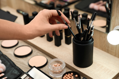 Professional makeup artist taking brush from holder at wooden table indoors, closeup