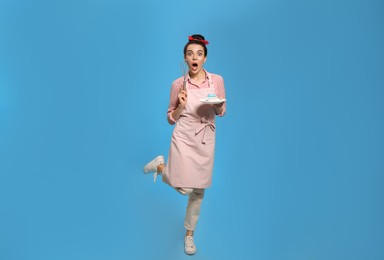 Emotional housewife with tasty cupcake and whisk on light blue background