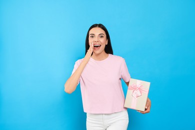 Emotional young woman holding gift box on light blue background