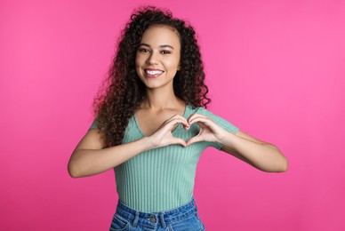 Happy young African-American woman making heart with hands on pink background
