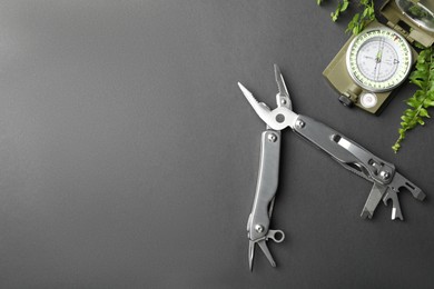 Compact portable metallic multitool, compass and green leaves on grey background, flat lay. Space for text