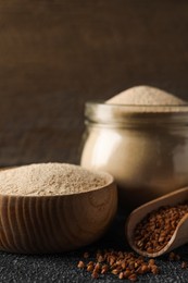 Bowl of buckwheat flour and scoop with grains on black table against wooden wall, closeup