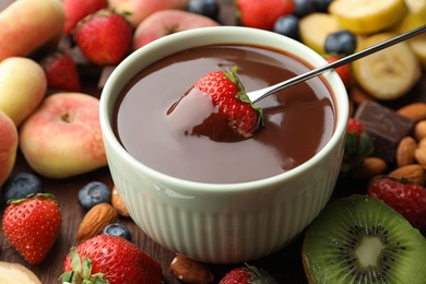 Photo of Fondue fork with strawberry in bowl of melted chocolate surrounded by other fruits on wooden table