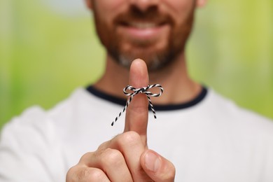 Man showing index finger with tied bow as reminder against green blurred background, focus on hand