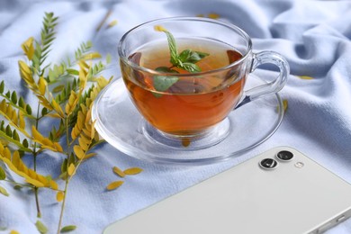 Cup of aromatic herb tea, smartphone and dry autumn leaves on white cloth
