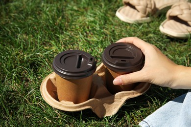 Woman holding takeaway cardboard coffee cups with plastic lids on green grass, closeup