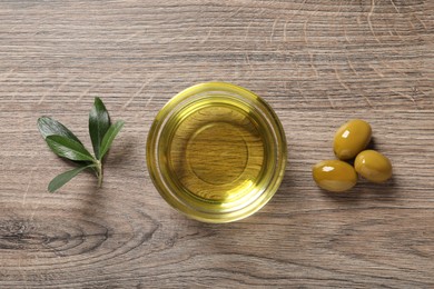 Glass bowl of oil, ripe olives and green leaves on wooden table, flat lay