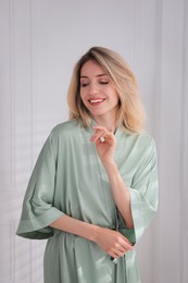 Pretty young woman in beautiful light green silk robe near white wall indoors
