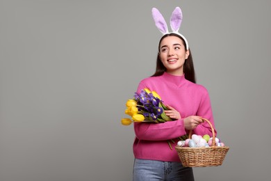 Photo of Happy woman in bunny ears headband holding wicker basket with painted Easter eggs and bouquet of flowers on grey background. Space for text