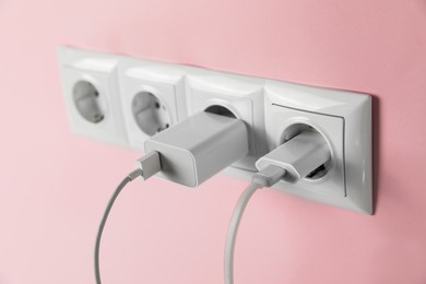 Charger adapters plugged into power sockets on pink wall, closeup. Electrical supply