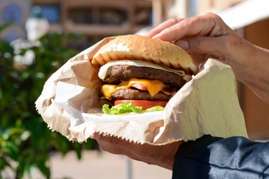 Woman holding delicious burger in paper wrap outdoors, closeup
