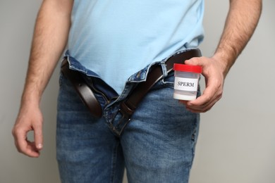 Donor with unzipped pants holding container of sperm on beige background, closeup