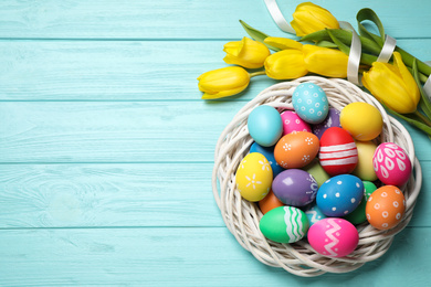 Colorful Easter eggs in decorative nest and tulip flowers on light blue wooden background, flat lay. Space for text