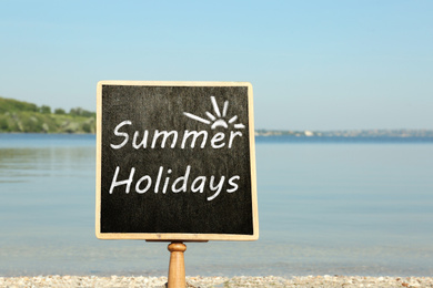 Image of Small chalkboard with inscription Summer Holidays on beach near river. School's Out