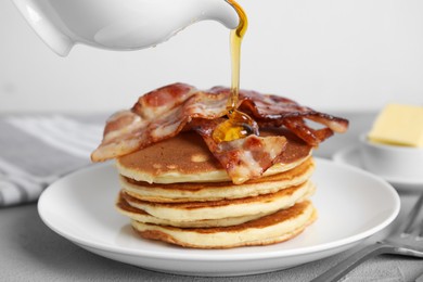 Pouring maple syrup onto delicious pancakes with fried bacon at grey table, closeup