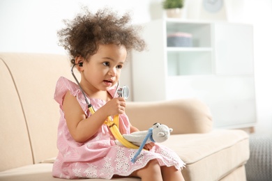 Cute African American child imagining herself as doctor while playing with stethoscope and toy on couch at home