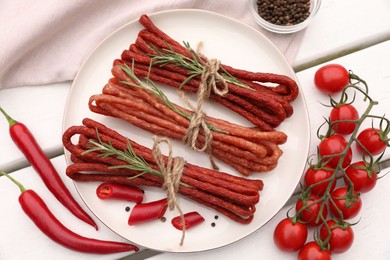 Photo of Bundles of delicious kabanosy with rosemary, peppercorn, chilli and tomatoes on white wooden table, flat lay