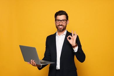 Photo of Smiling bearded man holding laptop and doing ok gesture on orange background