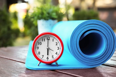 Photo of Alarm clock and fitness mat on wooden table outdoors. Morning exercise