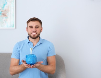 Young man with piggy bank on sofa in living room. Space for text