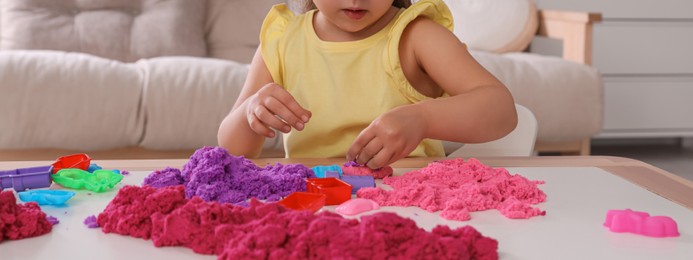 Cute little girl playing with bright kinetic sand at table in room, closeup. Banner design