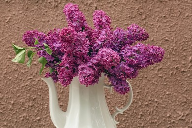 Beautiful lilac flowers in teapot near brown wall