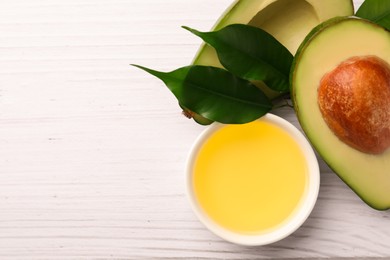 Photo of Cooking oil in bowl and fresh avocados on white wooden table, flat lay. Space for text