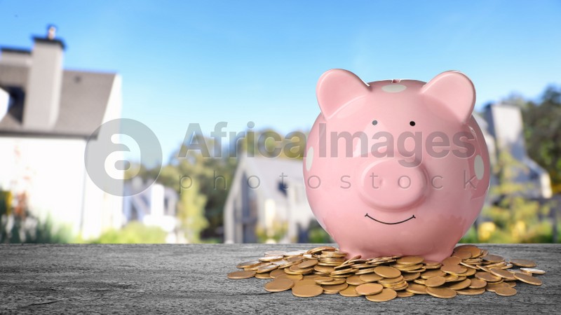 Piggy bank and coins on stone surface and blurred view of beautiful houses, space for text. Mortgage concept