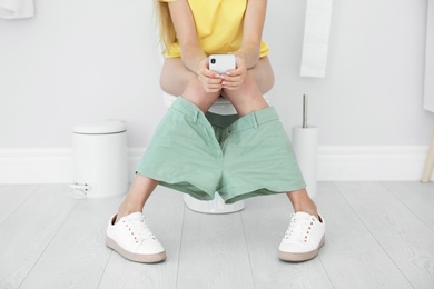 Young woman using mobile phone while sitting on toilet bowl at home