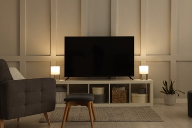 Photo of Modern TV on cabinet, armchair and lamps indoors. Interior design