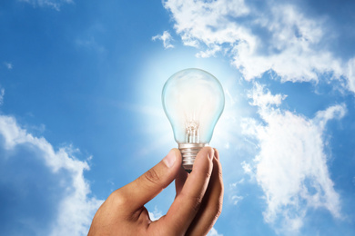 Solar energy concept. Man holding glowing light bulb against blue sky with clouds, closeup
