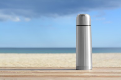 Metallic thermos with hot drink on wooden surface near sea, space for text