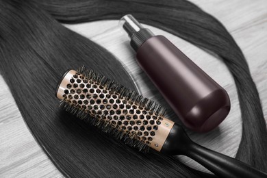 Photo of Spray bottle with thermal protection, lock of brunette hair and hairbrush on white wooden table, closeup