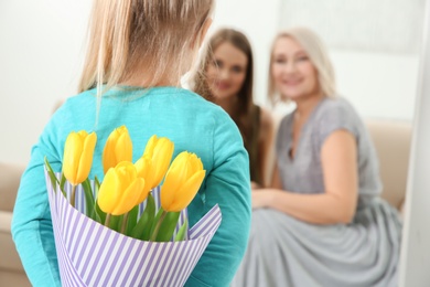 Little girl hiding tulip bouquet for mother and grandmother behind her back at home