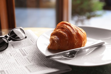 Tasty croissant, newspaper and sunglasses on table in cafeteria