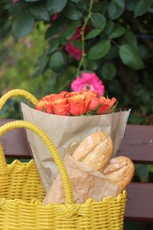 Photo of Beautiful bouquet of roses and baguettes in yellow wicker bag on bench outdoors