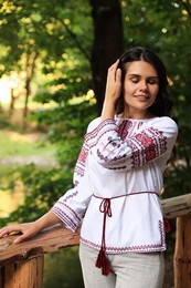 Beautiful woman wearing embroidered shirt near wooden railing in countryside. Ukrainian national clothes