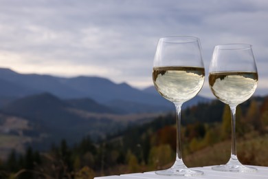 Photo of Glasses of tasty wine on white wooden table against mountain landscape. Space for text