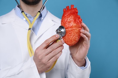 Doctor with stethoscope and model of heart on light blue background, closeup. Cardiology concept