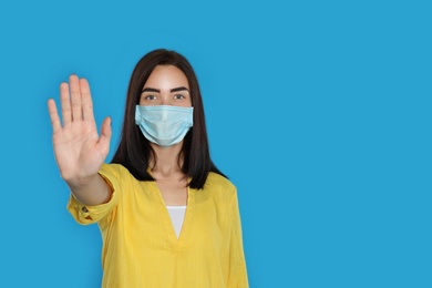 Young woman in protective mask showing stop gesture on light blue background, space for text. Prevent spreading of coronavirus