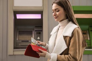 Photo of Woman putting money into wallet near cash machine outdoors
