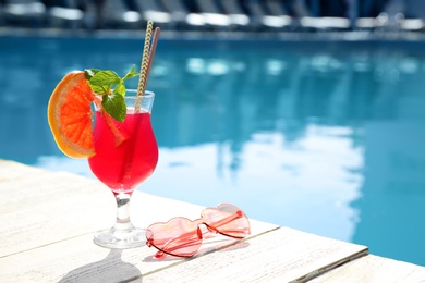 Refreshing cocktail and sunglasses near outdoor swimming pool on sunny day. Space for text
