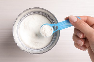 Woman taking powdered infant formula with scoop from can at white wooden table, top view
