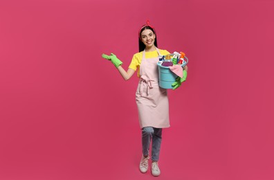 Young housewife holding bucket with cleaning supplies on pink background