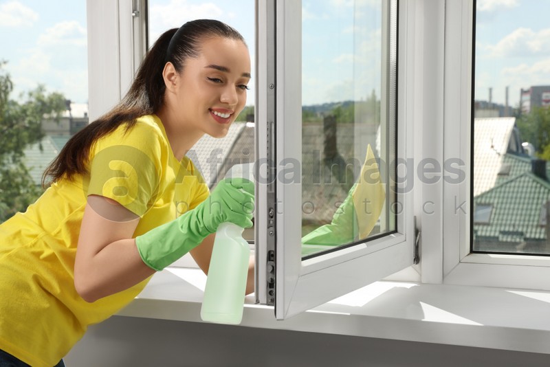 Happy young woman cleaning window glass with sponge cloth and spray indoors