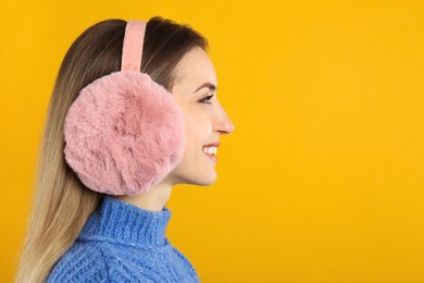 Happy woman wearing warm earmuffs on yellow background, space for text