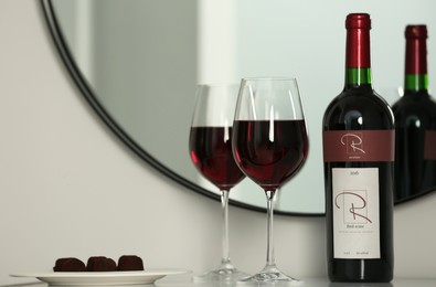 Bottle and glasses of red wine with chocolate candies on table near mirror